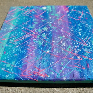 Könnte beinhalten: Abstraktes Gemälde auf Leinwand mit blauem, violettem und grünem Hintergrund. Das Gemälde zeigt weiße, rosa und blaue Spritzer und Linien.