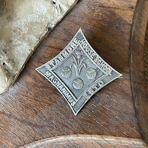 Può includere: Spilla argentata a forma di diamante con le parole "STANMORE HOUSE SCHOOL" e "MAGNANIMUS ESTO" incise sui bordi. Il centro presenta un motivo floreale con tre baccelli stilizzati.