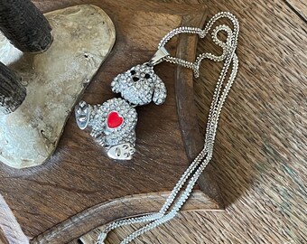 Può includere: Collana color argento con un ciondolo a forma di cane tempestato di cristalli trasparenti. Il cane ha un cuore rosso sul corpo. La collana è esposta su una superficie di legno.