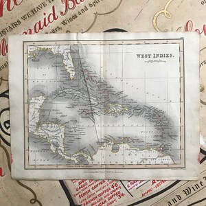 Op de afbeelding: Een antieke kaart van de West-Indische Eilanden, gedrukt in zwart-wit met rode accenten. De kaart toont de eilanden van de Caraïbische Zee, waaronder Cuba, Hispaniola, Puerto Rico en Jamaica. De kaart is getiteld "West Indies" en bevat een schaalbalk en een kompasroos.