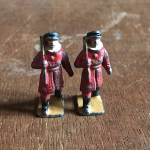 May include: Two miniature toy soldiers in red and black uniforms, holding rifles, standing on yellow bases.