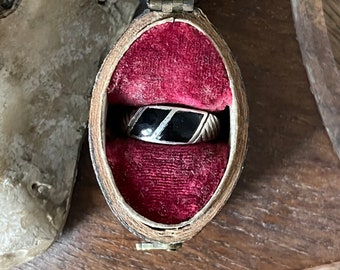 Könnte beinhalten: Ein silberner Ring mit schwarzem Inlay, präsentiert in einer ovalen Vintage-Schmuckschatulle mit rotem Samt. Der Ring hat ein geometrisches Design mit diagonalen Linien. Die Schatulle ist geöffnet und zeigt den Ring.