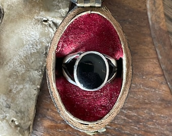 Könnte beinhalten: Ein silberner Ring mit einem schwarzen ovalen Edelstein, der in eine Ringfassung gefasst ist. Der Ring wird in einer roten Samtringschatulle präsentiert.