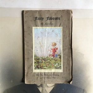以下が含まれることがあります： 茶色の背景に、妖精を見上げる女の子の絵が描かれた、ビンテージな本の表紙。本のタイトルは「Fairy Favours: An Anthology」で、絵の下には「With Best Wishes for Christmas and the Coming Year」というテキストが印刷されています。