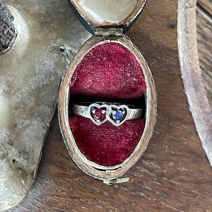 Peut inclure: Une bague en argent avec deux sertissages en forme de cœur entrelacés, l'un contenant une pierre précieuse rouge et l'autre une bleue. L'anneau est gravé des prénoms Natalie et Jess, présenté dans un écrin doublé de velours rouge.