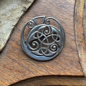 May include: A silver Celtic knot brooch with a circular design. The knot is intricate and detailed, with a smooth, polished finish.