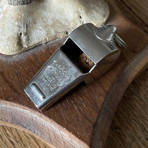 May include: A silver-colored Acme Thunderer whistle with a key ring, resting on a wooden surface. The text "THE ACME THUNDERER MADE IN ENGLAND" is visible on the whistle. The whistle is open, revealing a cork inside.