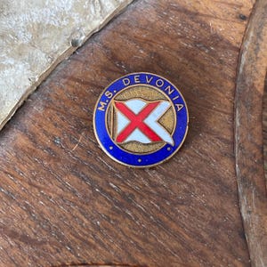 May include: A circular metal pin featuring a blue enamel border with the text M.S. DEVONIA in gold lettering. The center displays a white flag with a red cross design set against a textured gold background, enclosed by a thin gold metal rim.