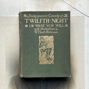 Pode incluir: Um livro verde com letras douradas intitulado "Shakespeare's Comedy of Twelfth Night or What You Will" com ilustrações de W. Heath Robinson. A capa apresenta uma ilustração dourada de um bufão tocando um alaúde.