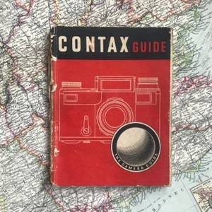 May include: A red book with the title "Contax Guide The Camera Guide" on the cover. The book features a black and white illustration of a Contax camera.