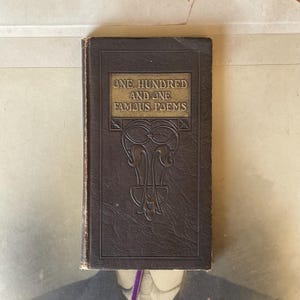 Puede incluir: Un libro antiguo de color marrón oscuro titulado "One Hundred and One Famous Poems". La portada presenta letras doradas y detalles decorativos en relieve. El libro parece desgastado, lo que sugiere que es una antigüedad o un objeto preciado.