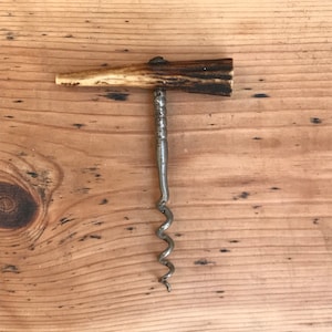 Könnte beinhalten: Antiker Korkenzieher mit einer Metallspirale und einem Griff aus hellbraunem und dunkelbraunem Geweih. Der Korkenzieher liegt auf einer hellbraunen Holzoberfläche.