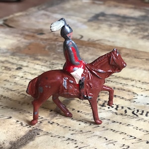 May include: A miniature toy soldier riding a brown horse. The soldier is wearing a red and white uniform with a gray helmet and a white feather. The horse has a brown saddle and bridle.