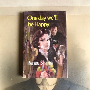 May include: A vintage paperback book cover with a painting of a woman in a black jacket and a bouquet of flowers. The title of the book is "One day we'll be Happy" by Renée Sharon.