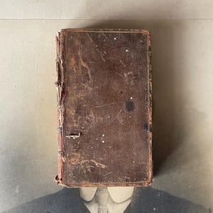 Può includere: Un libro antico, rilegato in pelle marrone usurata, con segni di usura visibili. La copertina mostra una texture invecchiata e una chiusura in metallo. Il dorso è visibile, rivelando la rilegatura. Probabilmente è un oggetto da collezione.