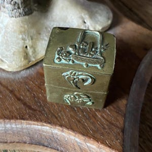 Puede incluir: Una pequeña caja antigua de latón con tapa con bisagras. La caja es cuadrada y presenta tallas en relieve detalladas de una cesta de flores y otros diseños florales. El latón tiene un aspecto deslustrado y envejecido, lo que sugiere su antigüedad.