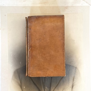 Puede incluir: Un libro antiguo con una cubierta de cuero marrón desgastado oscurece la cara de una persona con un traje gris. Las páginas del libro son visibles a lo largo del lomo, lo que sugiere edad y uso. El fondo es una fotografía descolorida.