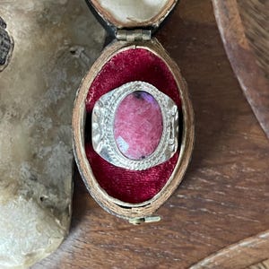 May include: A silver ring with an oval, faceted, pink gemstone. The ring is set in a decorative silver band with an ornate design. The ring is displayed in a vintage, velvet-lined jewelry box.