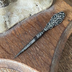 Könnte beinhalten: Eine antike silberfarbene Nähahle mit einem verzierten Griff mit floralem Design. Die Ahle hat eine lange, spitze Spitze und liegt auf einer Holzoberfläche.
