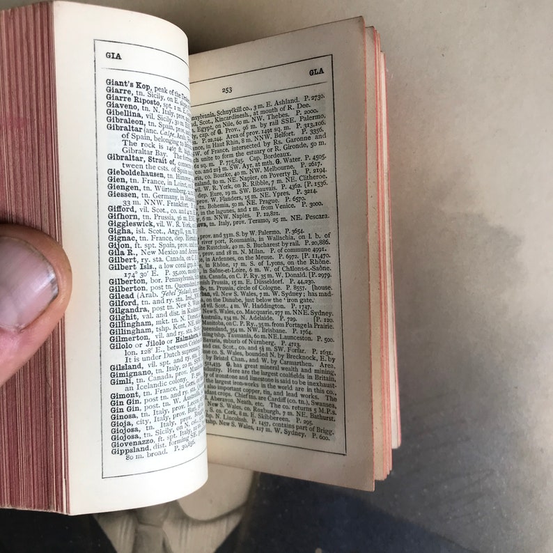 Op de afbeelding: Een open boek met een vergeelde pagina waarop een lijst met geografische locaties en hun bijbehorende beschrijvingen staat. Het boek is gebonden in bruin leer met gouden letters op de rug. De pagina is open op de letter "G" en bevat vermeldingen voor plaatsen zoals Gibraltar, Giarret, Giaveno en Giarre Ripostola.