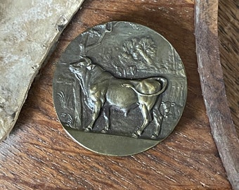 Puede incluir: Una medalla de bronce con una escultura en relieve detallada de un toro de pie en un campo. El toro está mirando hacia la izquierda y tiene un cuerno largo y curvo. La medalla es circular y tiene un borde elevado.