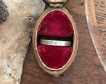Könnte beinhalten: Ein silberner Ring mit einem einzelnen Diamanten, der in einer quadratischen Form gefasst ist, liegt in einer kleinen, mit rotem Samt ausgekleideten Holzkiste.
