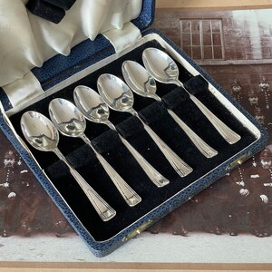 May include: A set of six silver teaspoons in a blue velvet lined box. The spoons are arranged in a row, with the handles facing the viewer. The box is closed, and the lid is slightly ajar.