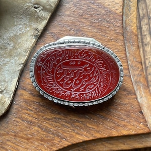Peut inclure: Une pierre de cornaline rouge avec une calligraphie arabe complexe gravée à la surface, sertie dans une bélière en argent avec un bord perlé.