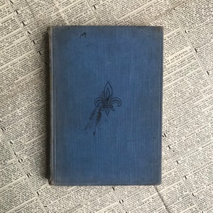 Può includere: Un libro rilegato in blu con un disegno a forma di giglio sulla copertina. Il libro è leggermente usurato e presenta alcune macchie scure sulla copertina.