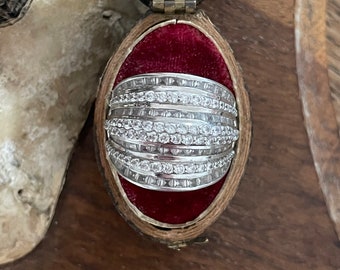 Könnte beinhalten: Ein silberner Ring mit mehreren Reihen kleiner, klarer Diamanten, die in einer Bandfassung gefasst sind. Der Ring wird in einer roten Samt-Ringbox präsentiert.