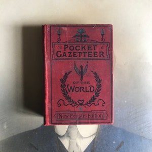 Op de afbeelding: Een rood hardback boek getiteld "Pocket Gazetteer of the World, New Census Edition". De omslag heeft een decoratief ontwerp met een gevleugeld wiel en een lauwerkrans.