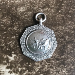 Peut inclure: Médaille en argent de forme octogonale représentant une scène de tir à la corde avec trois personnages. La médaille est ornée d'une bordure décorative et d'une boucle pour l'accrocher.