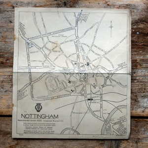 May include: A vintage map of Nottingham, showing roads, landmarks, and the AA logo. The map is printed in black ink on aged paper, with text indicating recommended and congested routes. The map is folded and placed on a wooden surface.