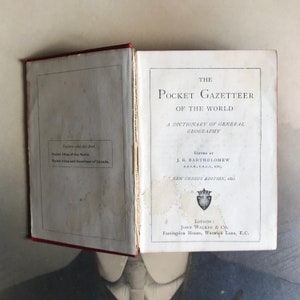 Op de afbeelding: Een vintage boek getiteld "The Pocket Gazetteer of the World: A Dictionary of General Geography", bewerkt door J.G. Bartholomew. De kaft van het boek is wit met zwarte tekst en een klein wapenschild in het midden. Het boek ligt open op de titelpagina.