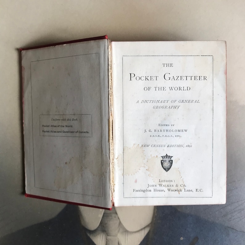 Op de afbeelding: Een vintage boek getiteld "The Pocket Gazetteer of the World: A Dictionary of General Geography", bewerkt door J.G. Bartholomew. De kaft van het boek is wit met zwarte tekst en een klein wapenschild in het midden. Het boek ligt open op de titelpagina.