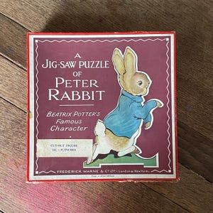May include: A red box with a white border and a cut-out image of Peter Rabbit. The box is a jigsaw puzzle of Beatrix Potter's famous character. The text on the box reads "A Jig-Saw Puzzle of Peter Rabbit, Beatrix Potter's Famous Character, Cut-Out Figure 14 1/2 x 9 1/2 inches, Frederick Warne & Co. Ltd. - London & New York. Made in Great Britain."