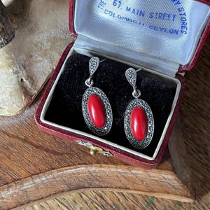 May include: Vintage silver earrings with oval red coral stones, framed by marcasite accents. The earrings are displayed in a red velvet jewelry box with text that reads "THE 67. MAIN STREET COLOMBO. CEYLON".