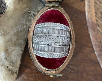 Könnte beinhalten: Ein silberner Ring mit mehreren Reihen von Baguette-Diamanten, die in einer runden Fassung gefasst sind. Der Ring wird in einer roten Samt-Ringbox präsentiert.