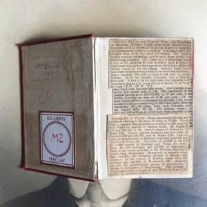 Op de afbeelding: Een close-up van een boek met een rode kaft en een boekenlegger met de tekst "EX LIBRIS M2 MACLAY". Het boek is open op een pagina met tekst over de hondenbelasting op het eiland Man en immoraliteit in Wales.