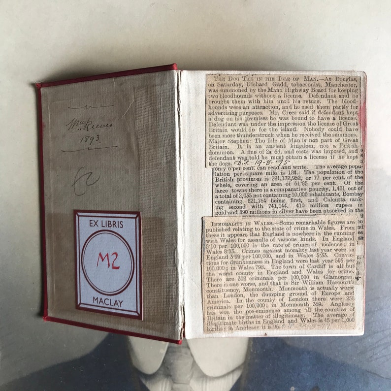 Op de afbeelding: Een close-up van een boek met een rode kaft en een boekenlegger met de tekst "EX LIBRIS M2 MACLAY". Het boek is open op een pagina met tekst over de hondenbelasting op het eiland Man en immoraliteit in Wales.