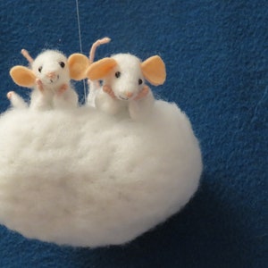 May include: Two white felt mice with pink ears are sitting on a white fluffy cloud. The cloud is hanging from a string against a blue background.