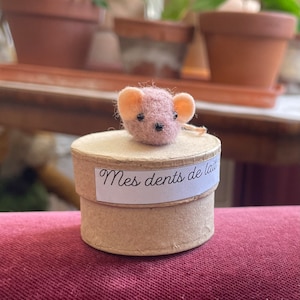 May include: A small pink felt mouse figurine sitting on top of a round cardboard box with the text "Mes dents de lait" written on a label.