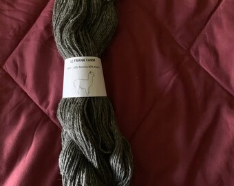 Puede incluir: Una madeja de hilo verde oscuro etiquetada "LC Frank Farm 20% Merino 80% Alpaca" con una pequeña ilustración de alpaca.