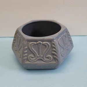 May include: A hexagonal ceramic planter with a matte periwinkle blue finish. The exterior features embossed scrollwork patterns on each side panel. The interior of the vessel is dark, and the rim is thick with a smooth, rounded edge design.