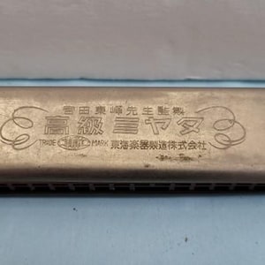May include: A silver-toned metal harmonica featuring engraved Japanese characters and decorative scrollwork on the top cover plate. The instrument has a rectangular body with visible air channels along the front edge and a metallic finish.