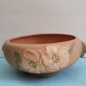 May include: A matte pink ceramic bowl featuring a raised relief design of two cream-colored flowers with pale green centers and leaves. The bowl has a rounded shape with small integrated handles on opposite sides and a smooth, uniform finish.