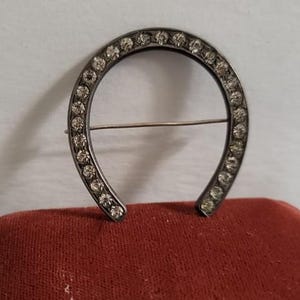 May include: A horseshoe-shaped brooch crafted from dark metal, featuring a series of clear, round-cut rhinestones embedded along the entire front surface. A thin, horizontal metal pin extends across the center of the open horseshoe frame.