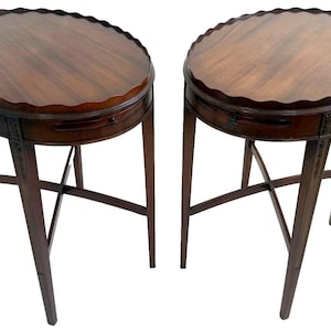 May include: Two antique mahogany side tables. Each table has a scalloped edge, a small drawer, and slender legs with cross supports. The tables are a rich, dark brown color and are set against a white background.