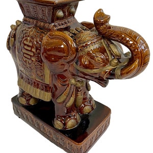 May include: A brown ceramic elephant garden stool with gold accents. The elephant has a raised trunk and a square top. The stool is decorated with intricate carvings.