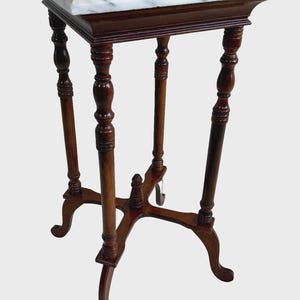 May include: A small, antique plant stand with a square marble top. The stand features a dark brown, wooden frame with turned legs and a decorative base. The marble top has gray veining.
